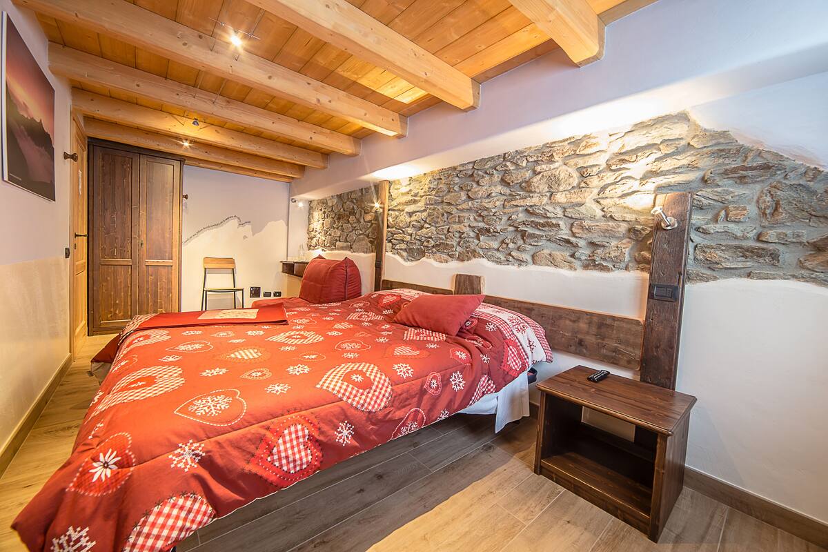 Une chambre avec un mur en pierre, des poutres apparentes en bois au plafond et un lit recouvert d’une courtepointe à motifs rouges.