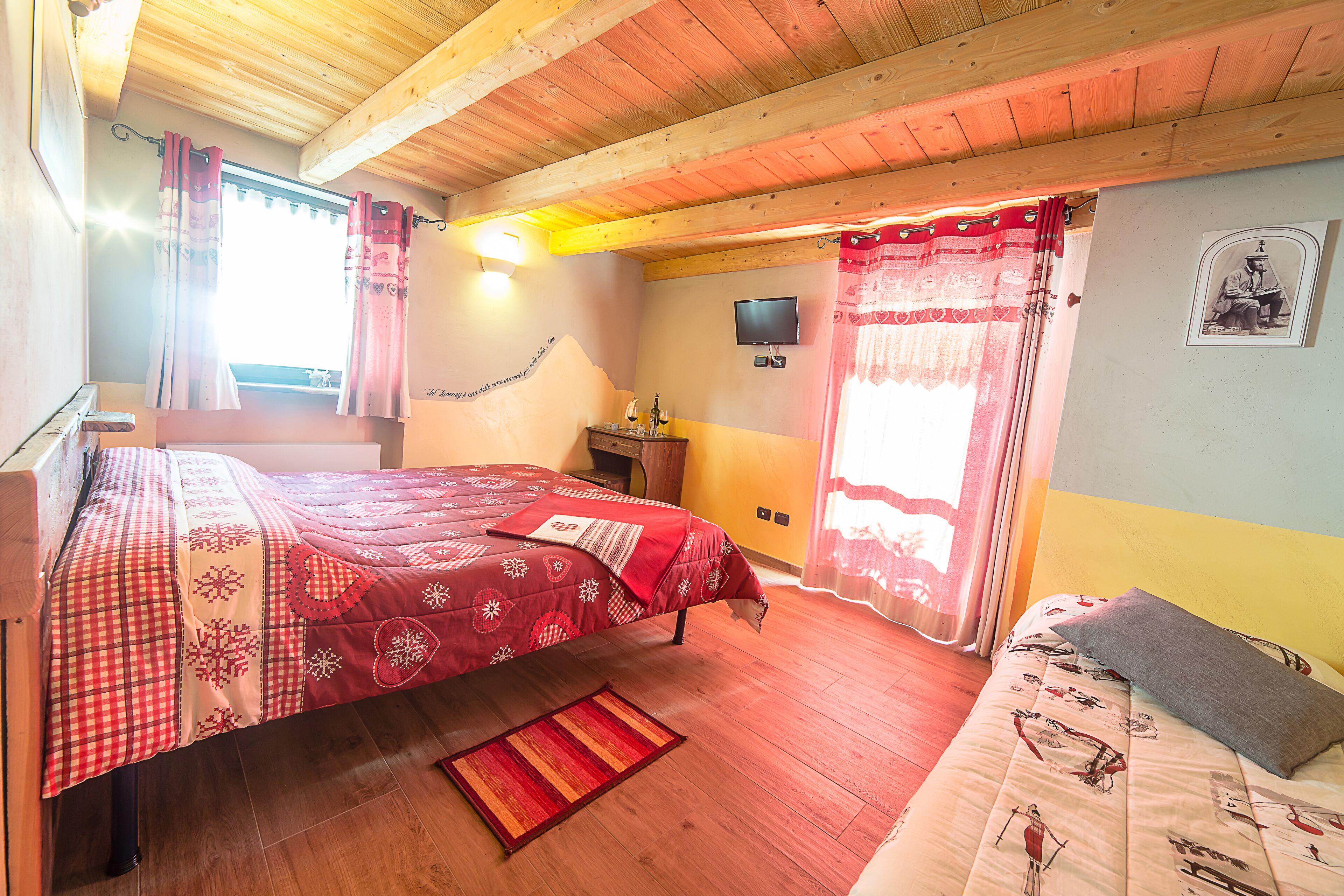 A bedroom with a wooden ceiling, a bed with a red and white patterned bedspread, a small bedside table, and a television on the wall.