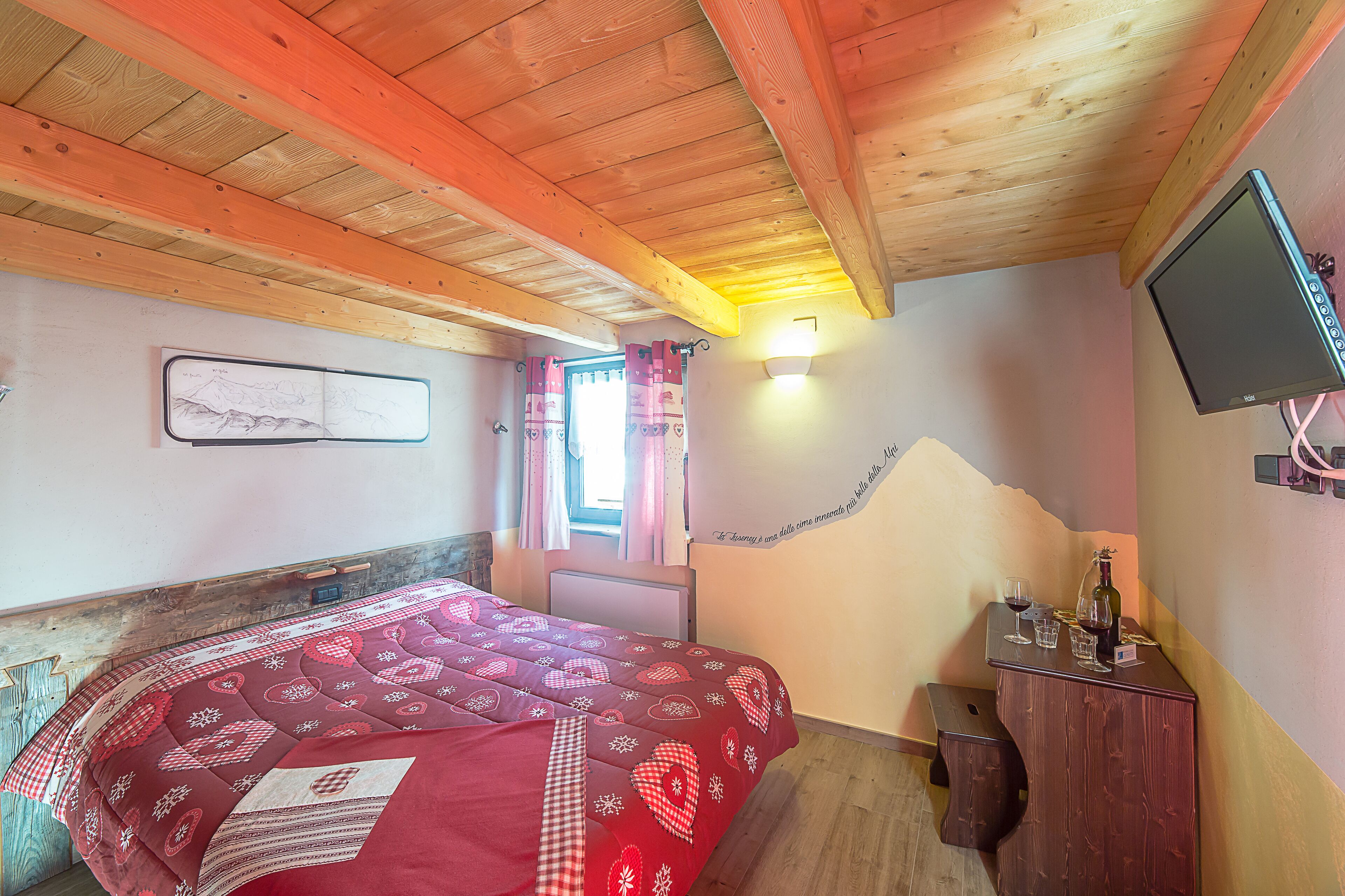 A bedroom with a wooden ceiling, a bed with a red quilt, a small table with a bottle and glasses, and a TV on the wall.