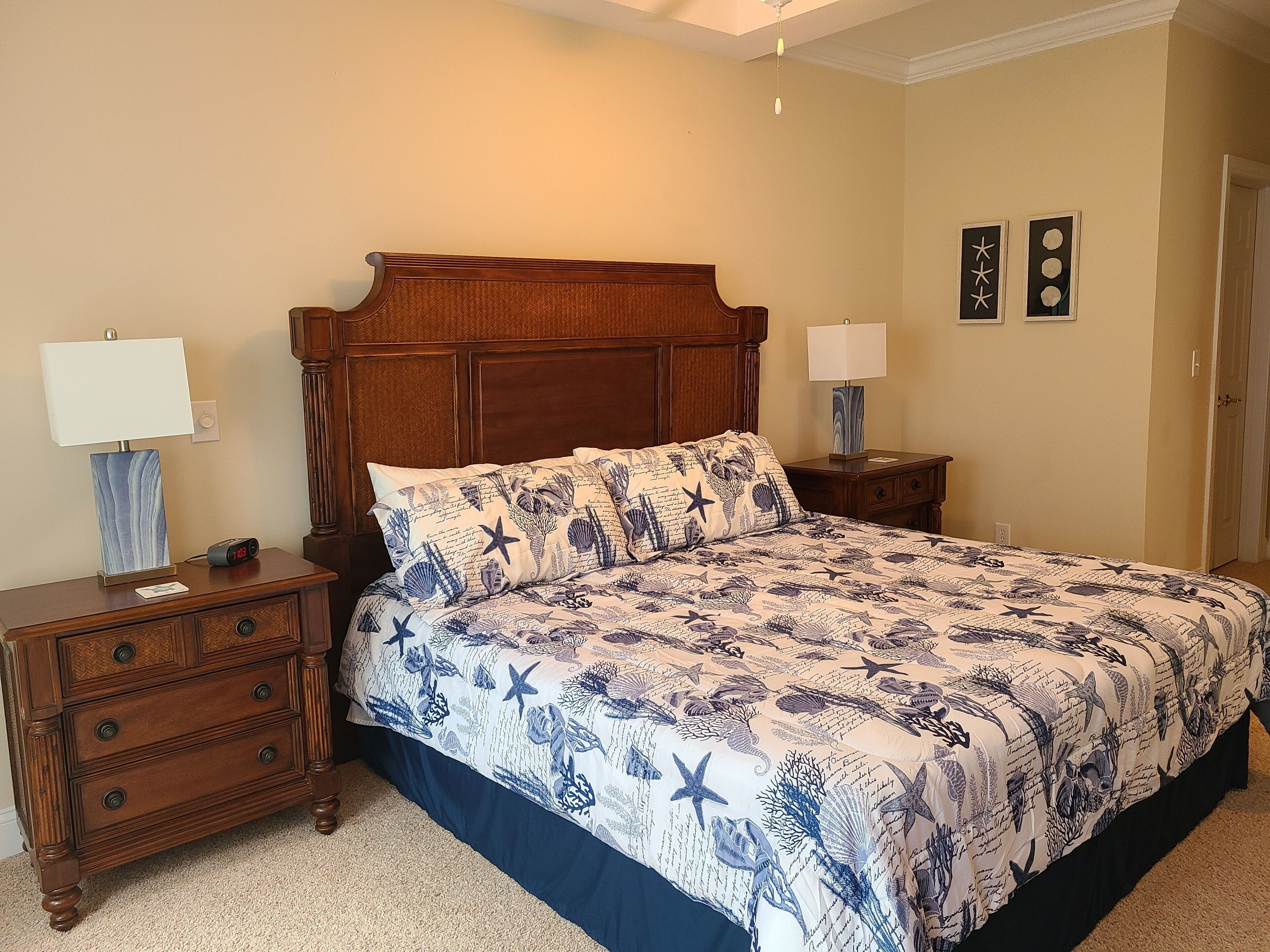 A bedroom with a wooden headboard, a bed with a blue floral bedspread, two bedside tables with lamps, and a nightstand with a digital clock.