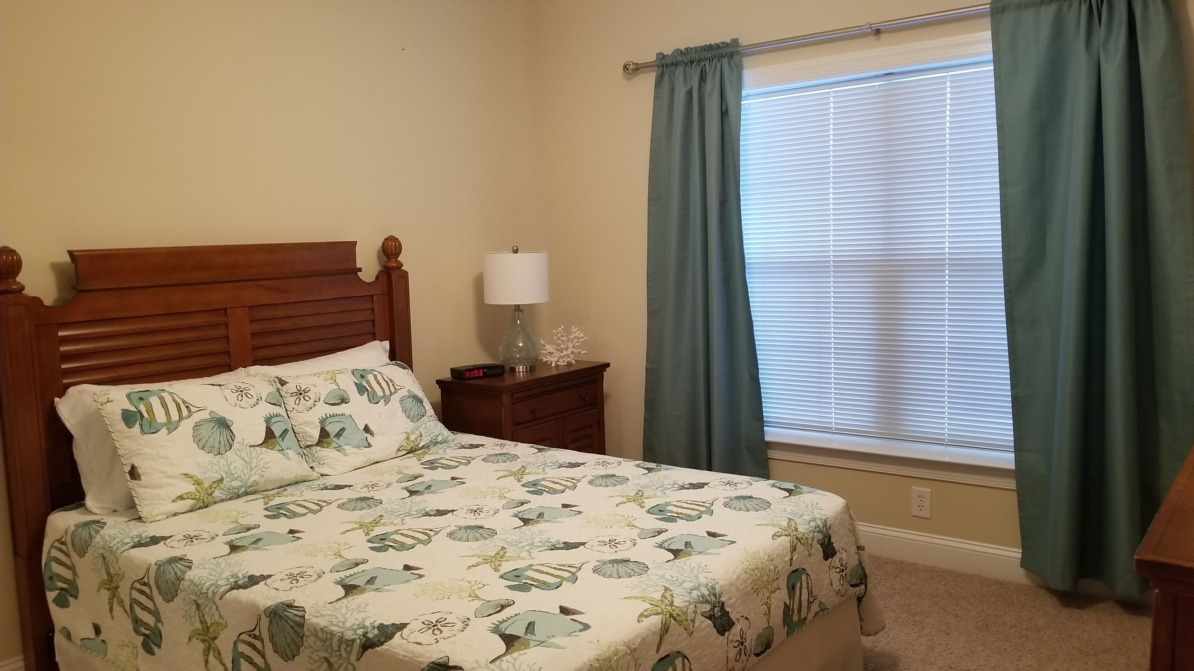 A bedroom with a wooden bed, a nightstand with a lamp, a clock, and a window with curtains.