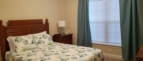 A bedroom with a wooden bed, a nightstand with a lamp, a clock, and a window with curtains.