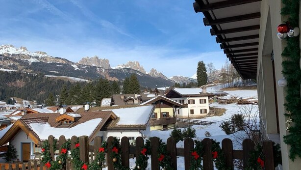 Ein Bergdorf mit traditionellen Chalets, einem Holzzaun, der mit Weihnachtsschmuck verziert ist, und einem klaren Blick auf schneebedeckte Berge im Hintergrund.