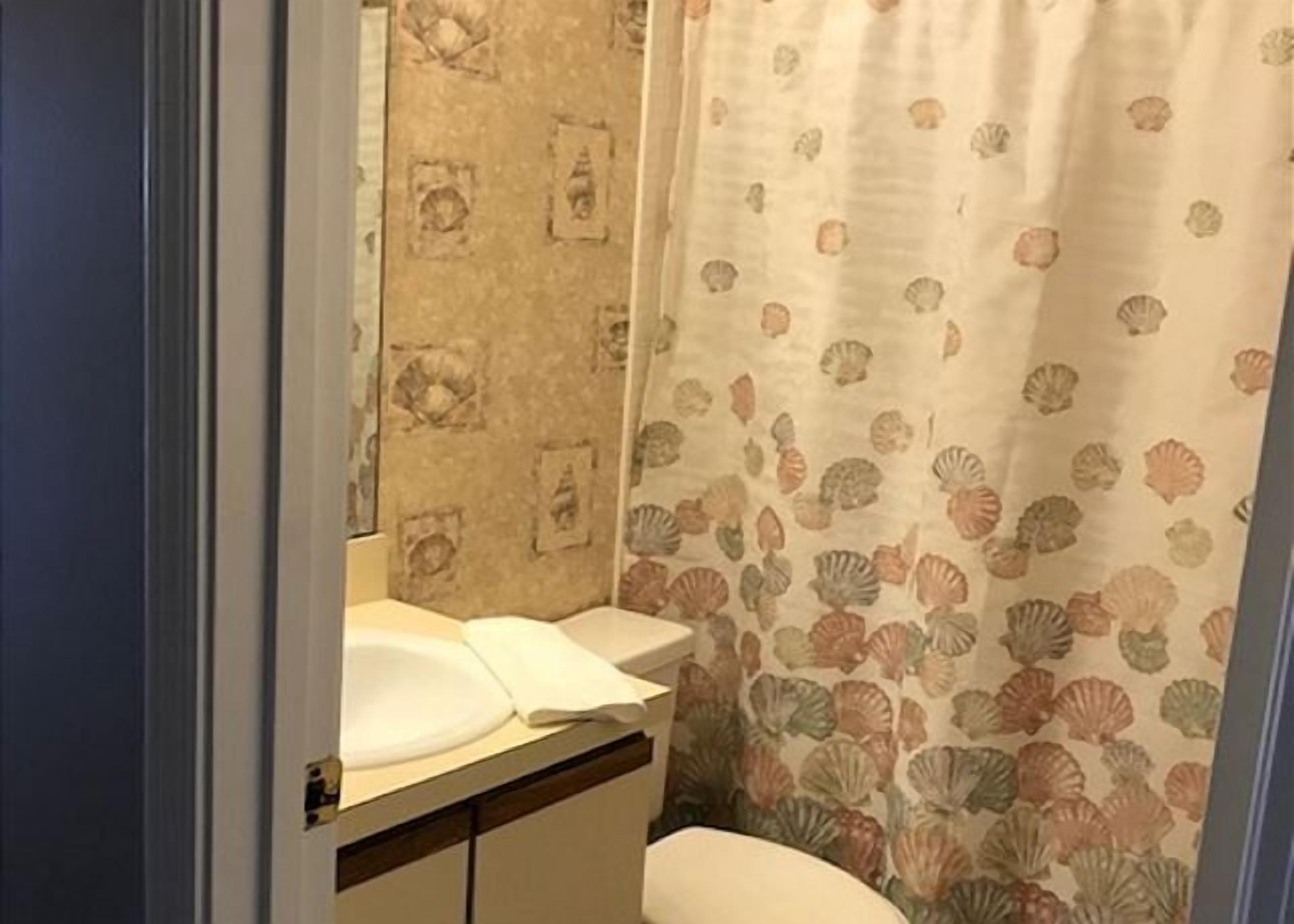 Une salle de bain équipée de toilettes, d’un lavabo et d’un rideau de douche orné d’un motif de coquillages.