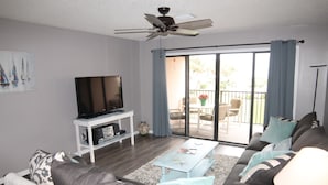A living room with a sofa, coffee table, TV, and a balcony with outdoor furniture. - Sea Place 13239 (St. Augustine)