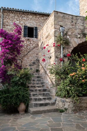 A stone staircase leading up to a building with a rustic stone facade, surrounded by lush greenery and vibrant flowers.