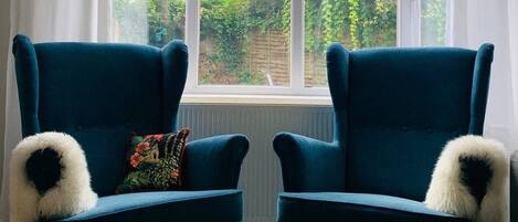 Two dark blue armchairs with fluffy white accents, a colorful floral pillow, and a wooden coffee table in front of a window with a view of a garden and buildings.