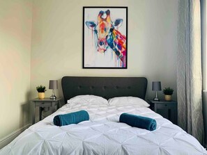 A neatly made bed with white bedding and two rolled towels, a framed abstract painting above the bed, and two bedside tables with lamps and small plants.