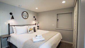 Una cama bien hecha con sábanas blancas, una mesita de noche con lámpara y una estantería en la pared con libros y un objeto decorativo.
