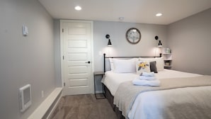 Ein Schlafzimmer mit weißer Tür, einem Bett mit weißer Bettwäsche, einem Nachttisch mit Lampe sowie einem an der Wand befestigten Regal mit Büchern und einer Pflanze.