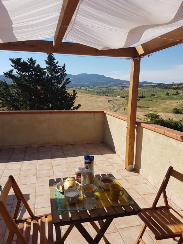 Un porche cubierto con una mesa lista para el desayuno, con vistas a un paisaje pintoresco con colinas y árboles.