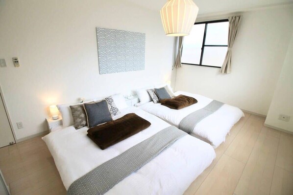 A hotel room with two beds, a bedside table with a lamp, a window with curtains, and a wall art piece.
