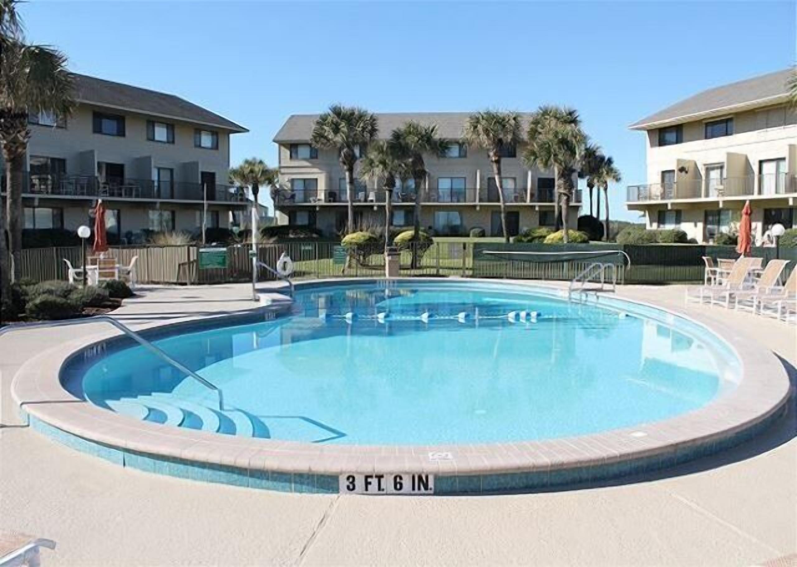A swimming pool with a 3 ft. 6 in. depth marker, surrounded by a fence, with palm trees and a building in the background.