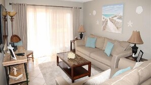 Un salon avec un canapé beige, une table basse en bois et une lampe. Il y a une fenêtre avec des rideaux et un tableau représentant une scène de plage accroché au mur.