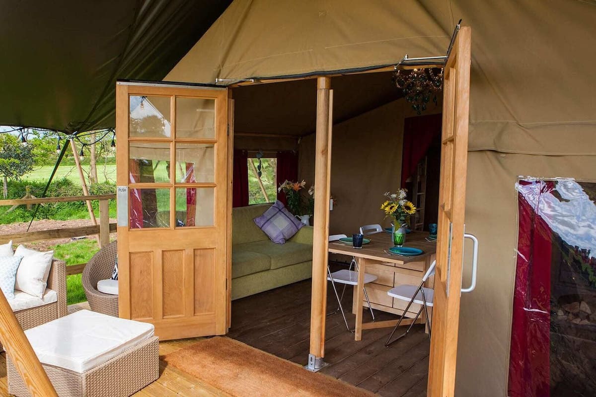 Een gezellig tent met een houten deur, een eethoek met tafel en stoelen, en een zithoek met een bank en kussens.
