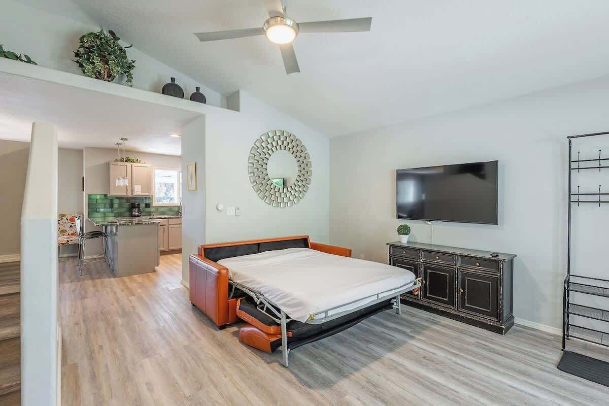 A bedroom with a bed, a TV, a ceiling fan, and a kitchen area visible through an open door.