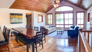 A spacious living area with a wooden dining table, sofa, and chairs, large windows, and a ceiling fan. - The Landing on Lake Nebagamon - Includes Pontoon! (Lake Nebagamon)