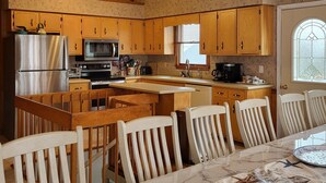 Eine Küche mit Holzschränken, einem Kühlschrank, einer Mikrowelle und einem Herd. Es gibt einen Esstisch mit Stühlen und ein Fenster mit Jalousien.