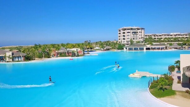 A large, clear blue swimming pool with a view of a resort building and palm trees in the background.