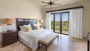 A bedroom with a bed, nightstands, a bench, a ceiling fan, and a view of greenery through large sliding doors.