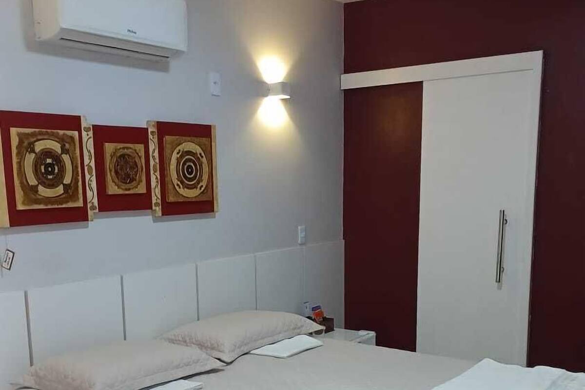 A neatly made bed with a white comforter and pillows, a wall-mounted air conditioner, and three decorative wall art pieces.