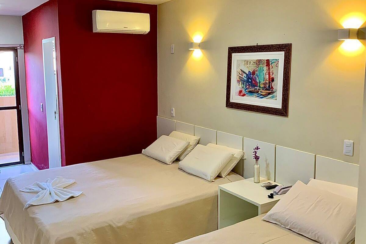 A hotel room with two beds, a red accent wall, an air conditioning unit, a framed artwork, and a bedside table with a vase.