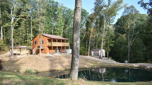 Eine zweistöckige Holzhütte mit Balkon, einem kleinen Schuppen und einem Teich in einem bewaldeten Gebiet.