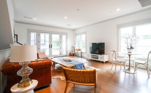 Amplia sala de estar con sofá de cuero, una mesa de centro de madera y un televisor sobre un mueble blanco. Amplias ventanas ofrecen luz natural y vistas al exterior.
