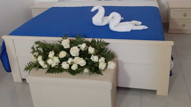 A bed with a blue bedspread and two white swan-shaped pillows. There is a white bedside table with a floral arrangement on the foot of the bed. - Giosy (Portopalo di Capo Passero)