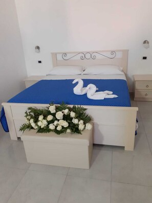 A bed with a blue bedspread and two white swan-shaped pillows. There is a white bedside table with a floral arrangement on the foot of the bed. - Giosy (Portopalo di Capo Passero)