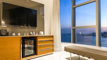 Habitación de hotel moderna con televisor grande, minibar y un banco junto a una ventana amplia con vistas a la ciudad.