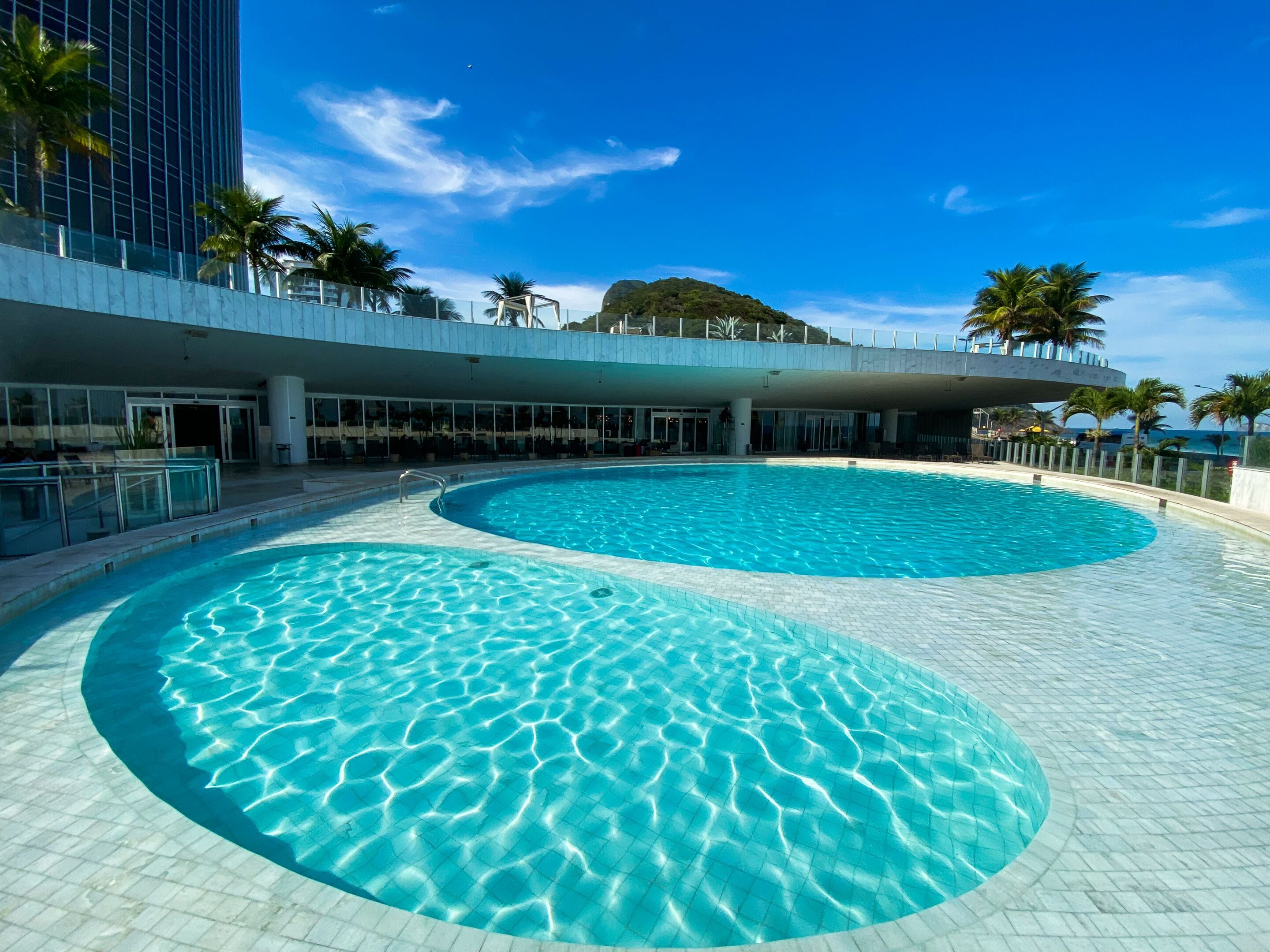 Uma grande piscina ao ar livre com um cenário arquitetônico moderno, incluindo um arranha-céu e palmeiras.