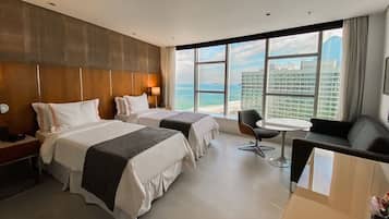 A hotel room with two beds, a sofa, a small table, and a view of the ocean and buildings.