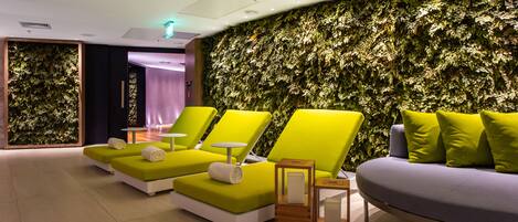 Un área de spa moderna con sillones reclinables verde y amarillo, un sofá púrpura y una pared con diseño de hojas.