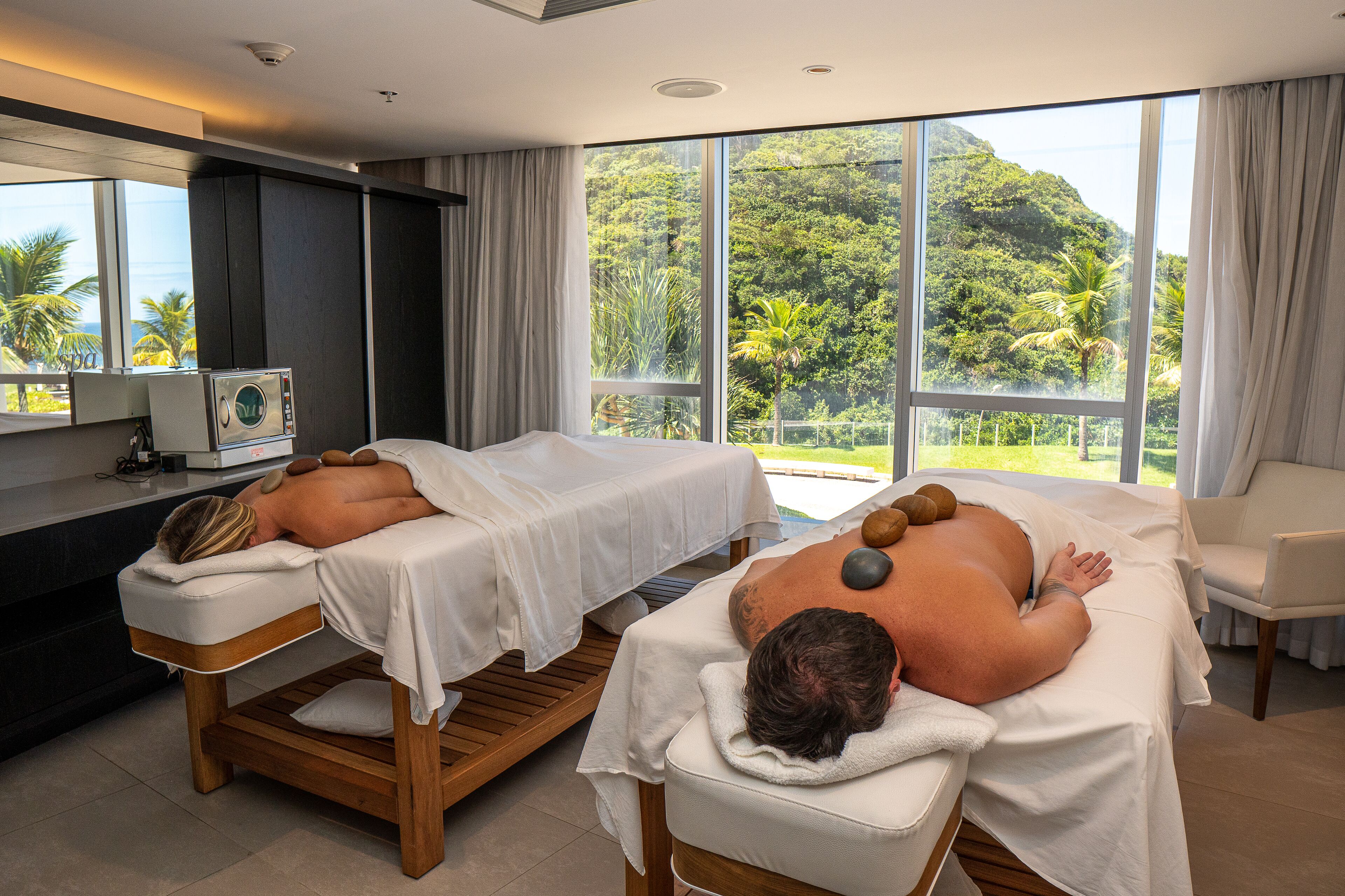 Un cuarto de spa con dos camillas de masaje, una silla de masaje y vistas a vegetación exuberante.