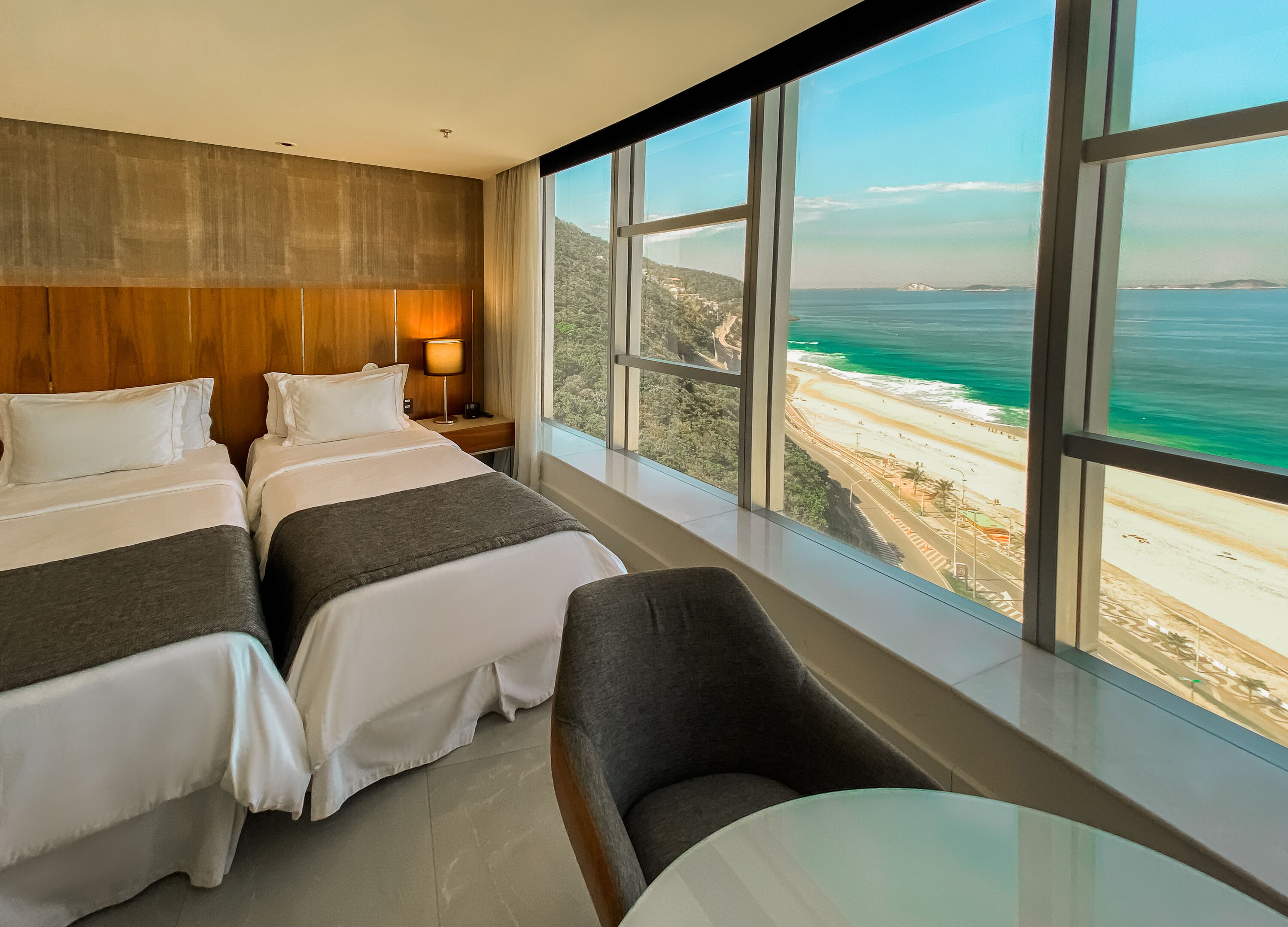 Habitación de hotel con dos camas, una mesita de noche, una mesa y vista a la playa y al océano.