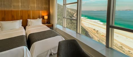 Habitación de hotel con dos camas, una mesita de noche, una mesa y vista a la playa y al océano.