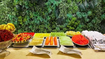 Uma mesa de buffet com diversas frutas e legumes, incluindo uvas, melão e kiwi, com um cenário de plantas artificiais ao fundo.