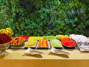 Una mesa de buffet con diversas frutas y verduras, como uvas, melón y kiwi, frente a un fondo de follaje artificial.