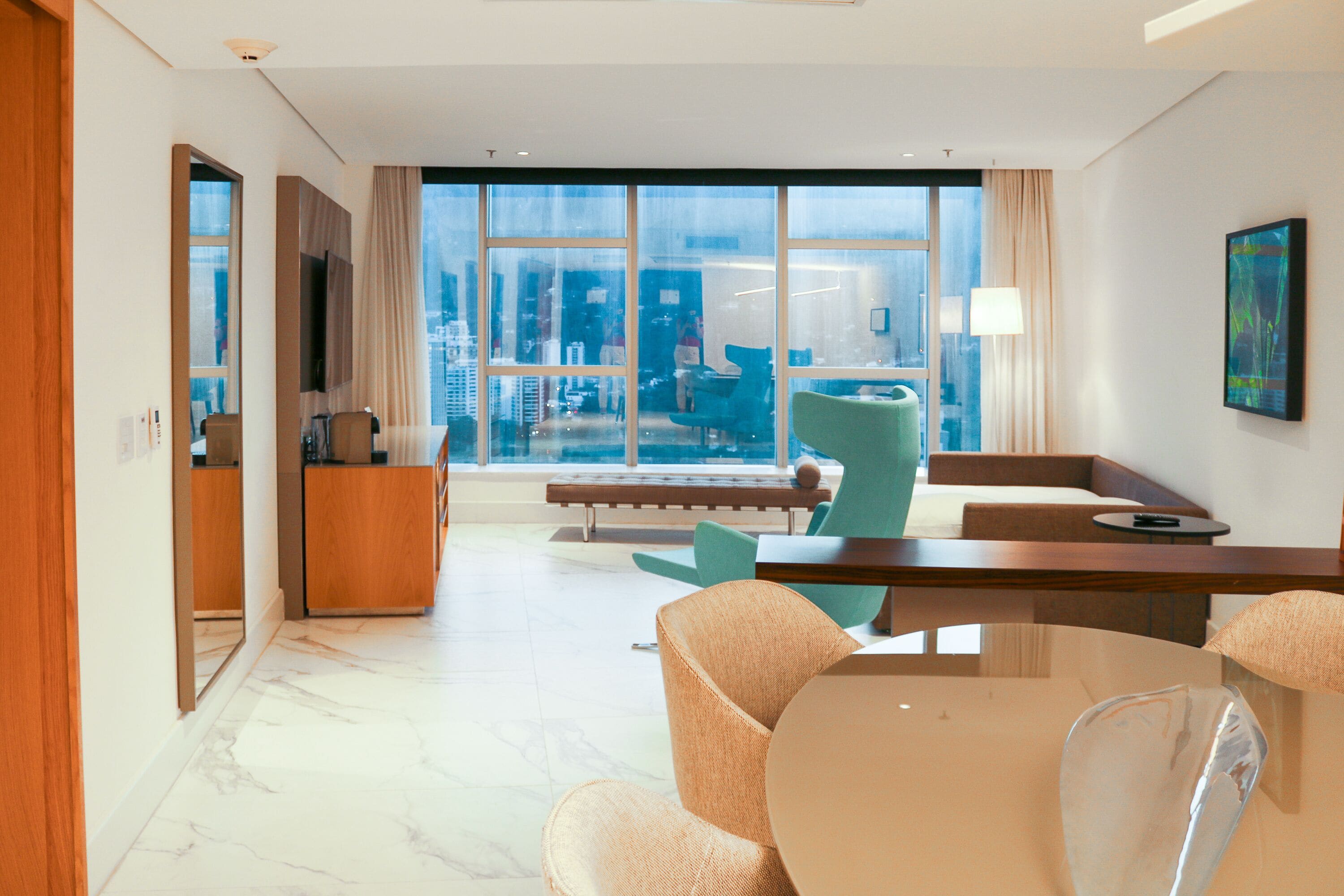 A modern hotel room with a large window, a dining area with a table and chairs, a sofa, and a television.