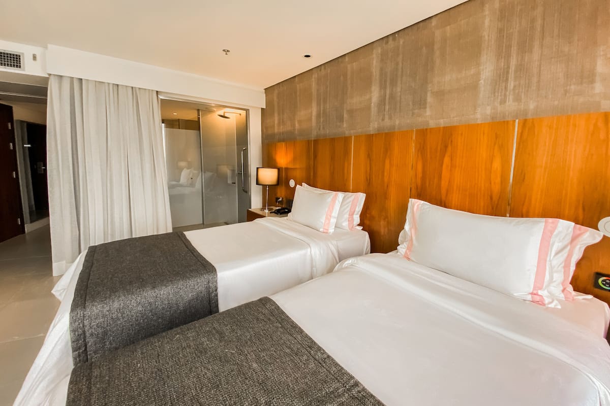 A hotel room with two beds, a wooden headboard, a nightstand with a lamp, and a bathroom visible through a glass door.