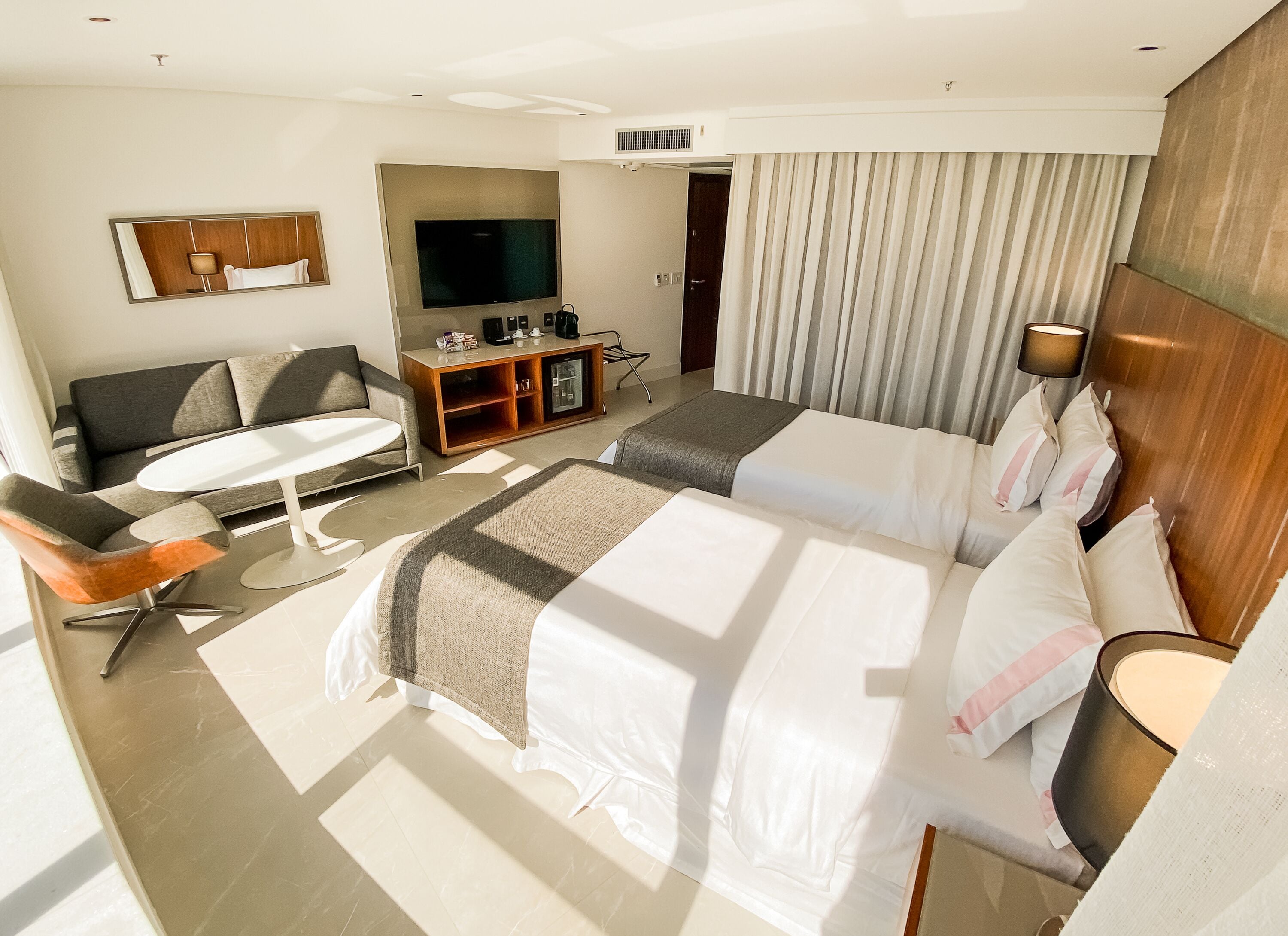 A hotel room with a large bed, a sofa, a small table, a TV, and a minibar.