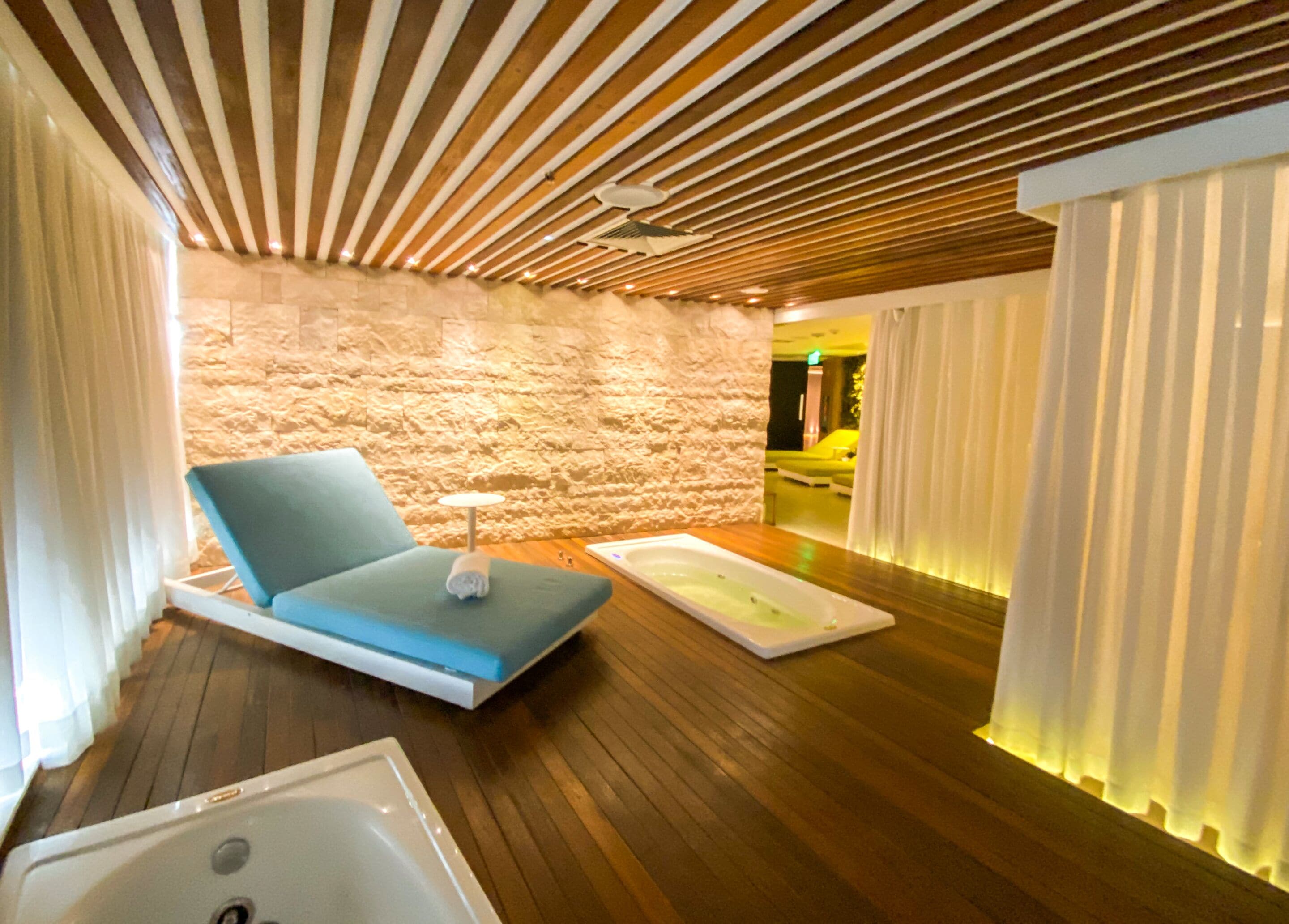 Un moderno spa con techo de madera, pared de piedra, una silla de descanso azul, una bañera blanca y una camilla de masaje.
