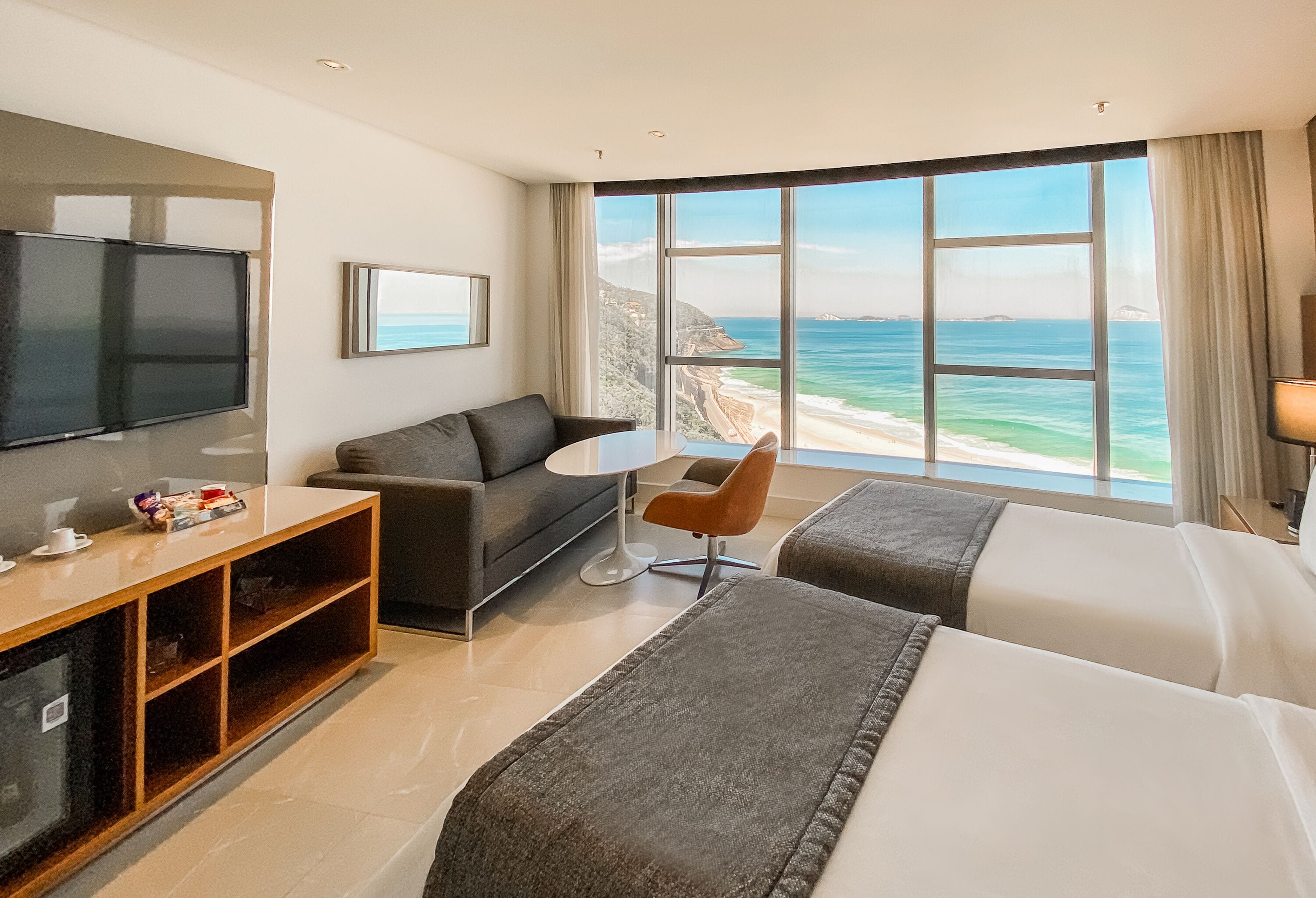 A hotel room with a large window offering a view of the ocean, a flat-screen TV, a sofa, a small table, and two beds.