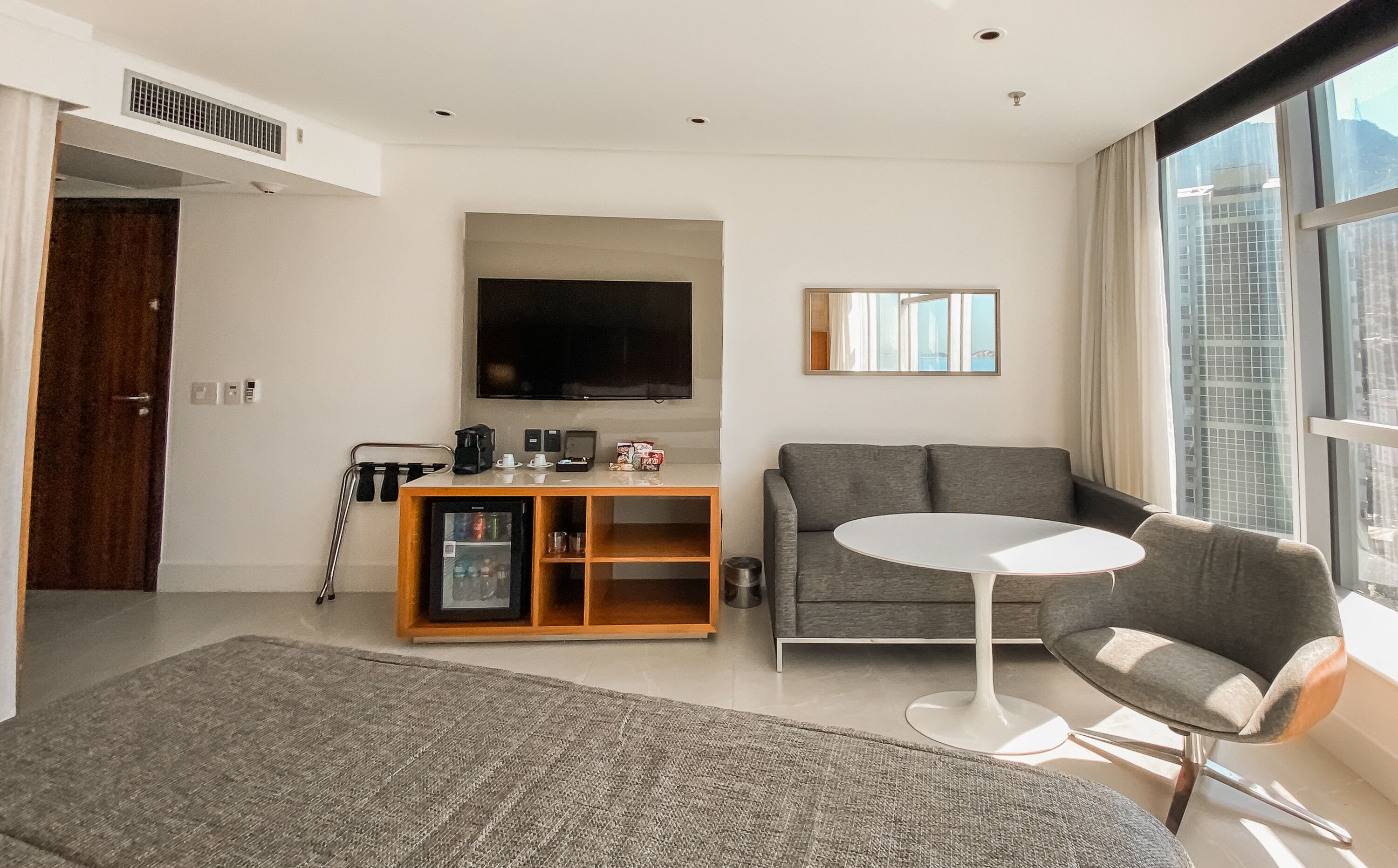 A modern living room with a grey sofa, a white coffee table, a TV, and a large window with a city view.