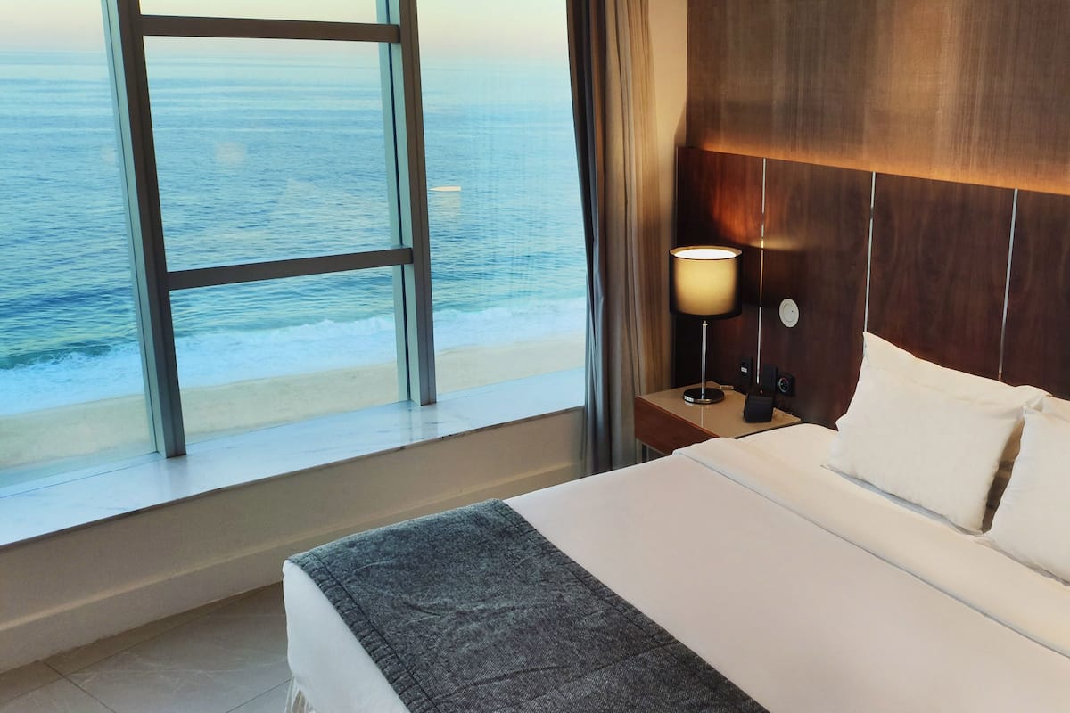 A hotel room with a large window offering a view of the ocean, a bed with white linens, a wooden headboard, and a bedside lamp.