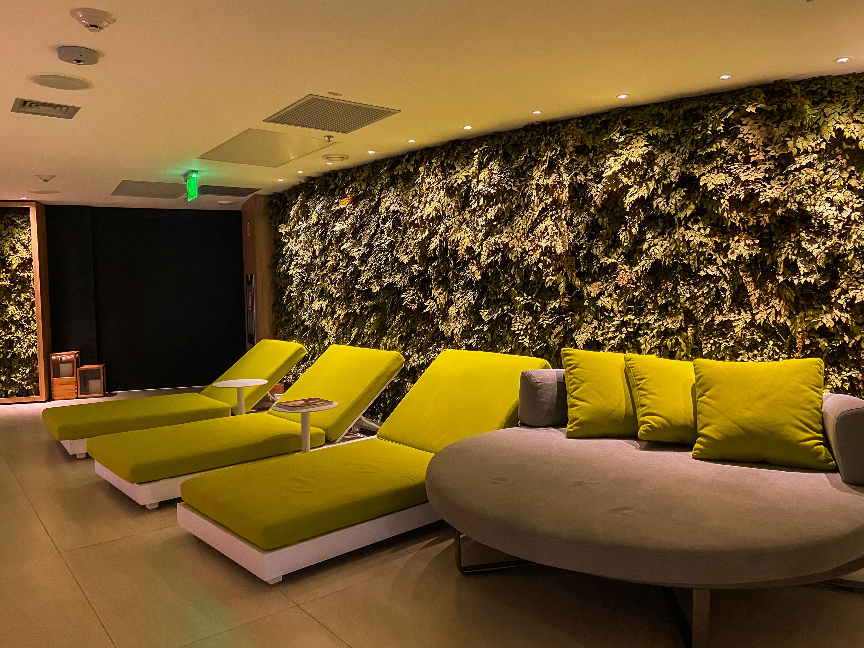 A modern lounge area with green and grey lounge chairs, a grey sofa, and a wall covered in foliage.