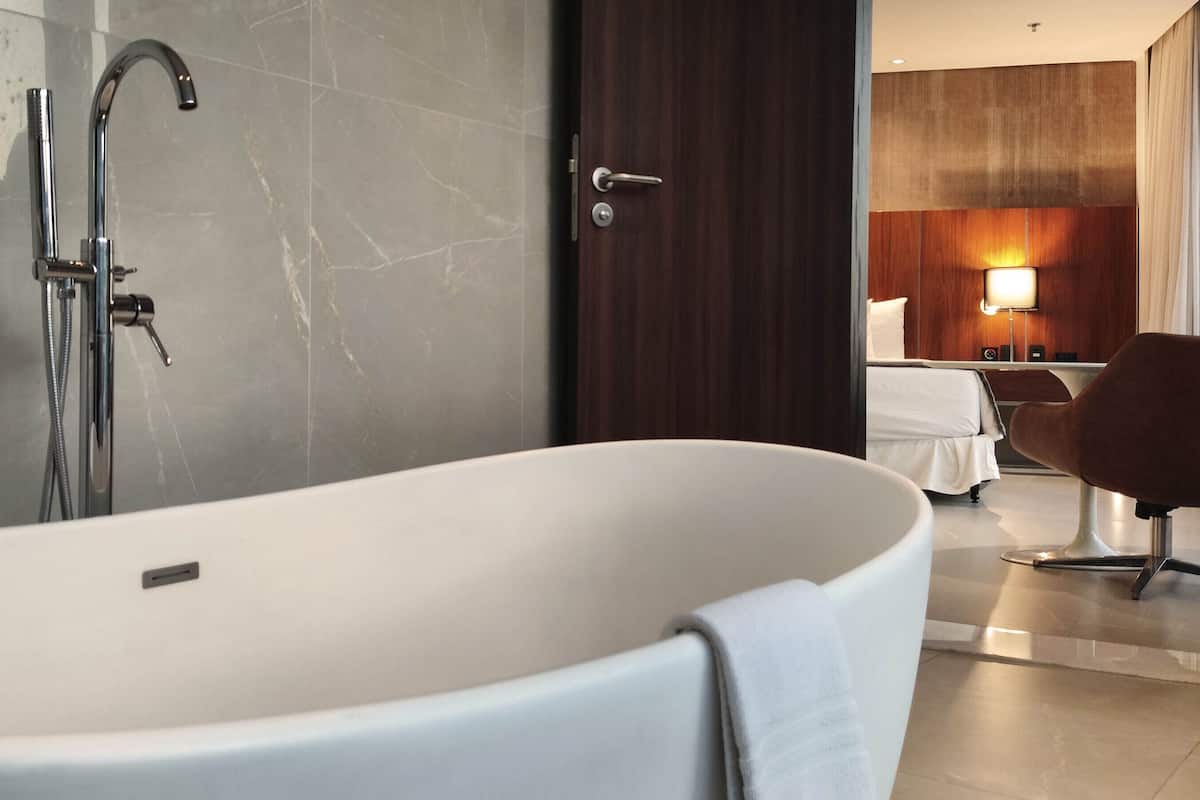 A modern bathroom with a freestanding bathtub, a showerhead, and a door leading to a bedroom with a bed and a chair.