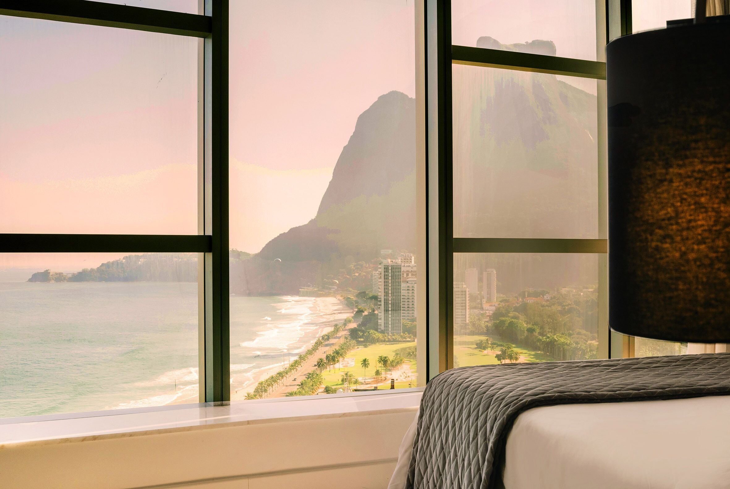 A hotel room with a large window offering a view of a beach, ocean, and a mountain.