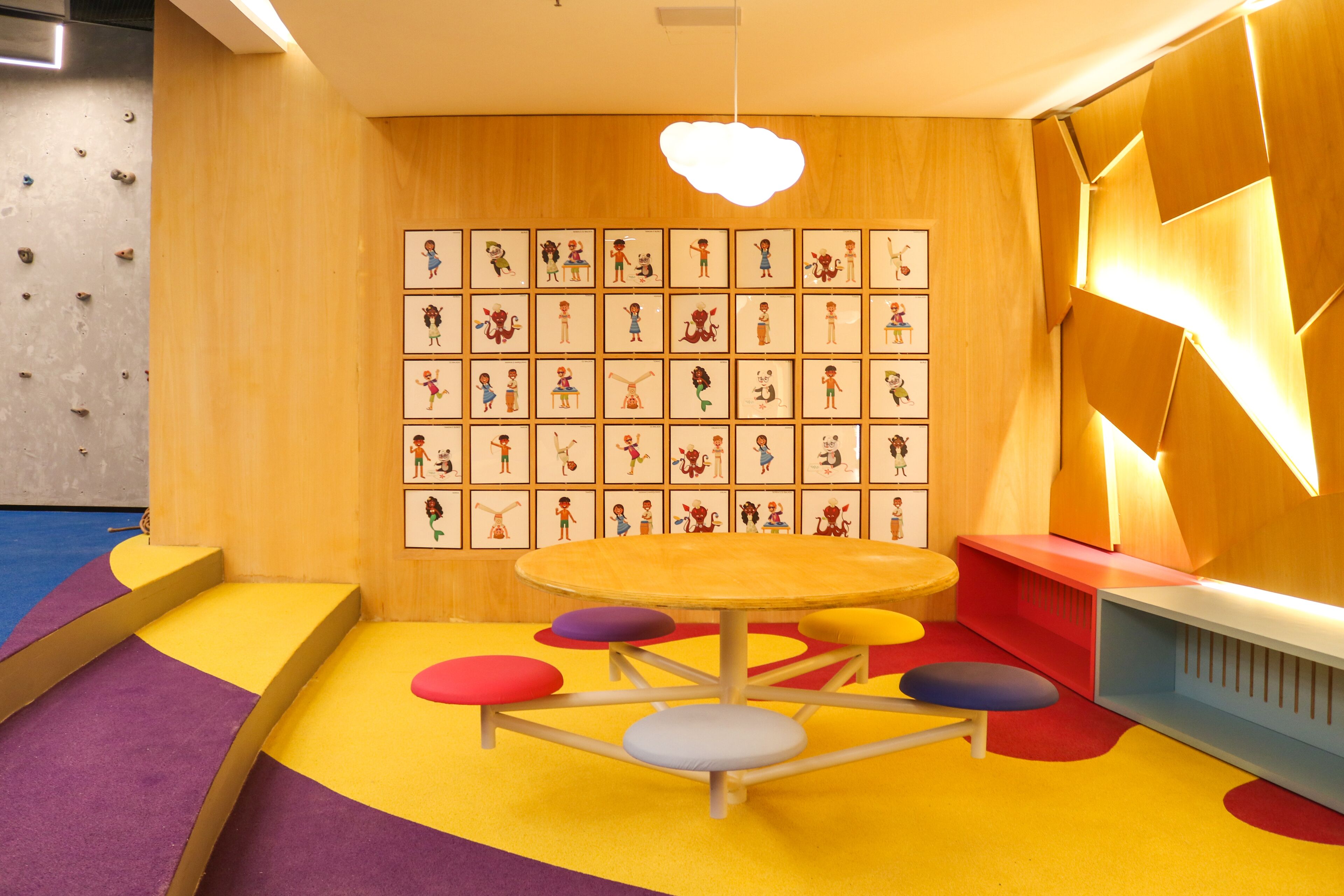 A playroom with a climbing wall, colorful play mats, a round table with stools, and a wall with framed illustrations.